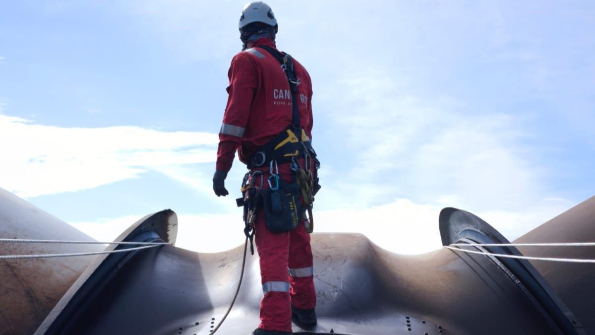 Técnico de trabajos verticales con equipo de protección individual completo, de pie sobre una gran estructura metálica industrial. Lleva casco, arnés y herramientas sujetas al cinturón mientras observa el entorno. Dos líneas de cuerda atraviesan la escena y el cielo despejado sirve de fondo, destacando un entorno de trabajo en altura y acceso mediante cuerdas.
